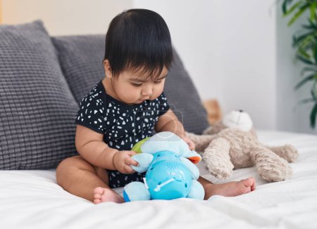Foto de Adorable bebé hispano jugando con muñecas sentadas en la cama en el dormitorio - Imagen libre de derechos