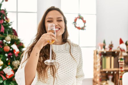 Téléchargez les photos : Jeune belle femme hispanique buvant du champagne debout près de l'arbre de Noël à la maison - en image libre de droit