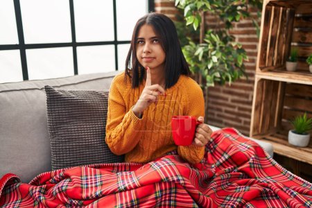 Foto de Young hispanic woman sitting on the sofa drinking a coffee at home thinking concentrated about doubt with finger on chin and looking up wondering - Imagen libre de derechos
