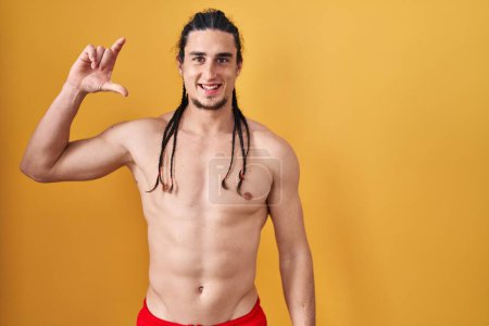 Photo for Hispanic man with long hair standing shirtless over yellow background smiling and confident gesturing with hand doing small size sign with fingers looking and the camera. measure concept. - Royalty Free Image