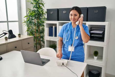 Photo for Young hispanic woman wearing doctor uniform talking on the telephone at clinic - Royalty Free Image