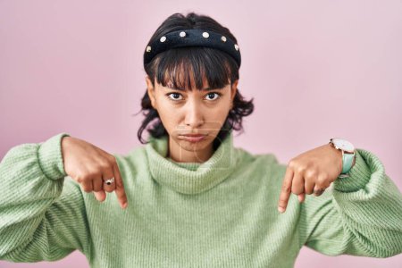 Foto de Young beautiful woman standing over pink background pointing down looking sad and upset, indicating direction with fingers, unhappy and depressed. - Imagen libre de derechos