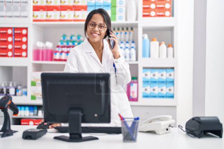 Photo for Young hispanic woman pharmacist talking on smartphone using computer at pharmacy - Royalty Free Image