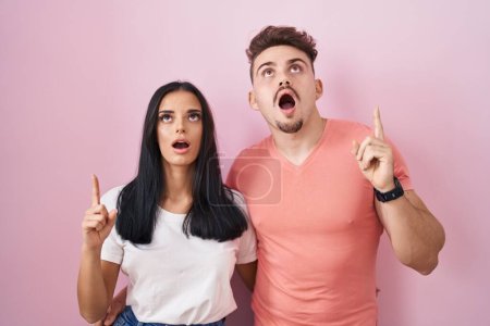 Téléchargez les photos : Jeune couple hispanique debout sur fond rose étonné et surpris regardant vers le haut et pointant avec les doigts et les bras levés. - en image libre de droit