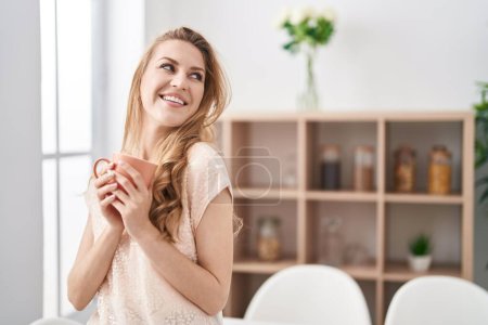Téléchargez les photos : Jeune femme blonde souriante confiance boire du café à la maison - en image libre de droit