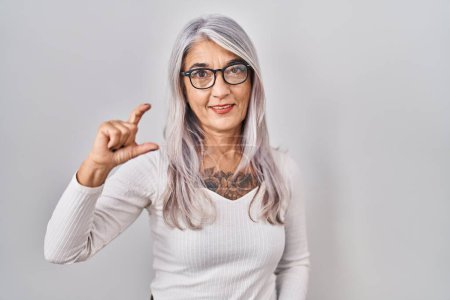 Téléchargez les photos : Middle age woman with grey hair standing over white background smiling and confident gesturing with hand doing small size sign with fingers looking and the camera. measure concept. - en image libre de droit