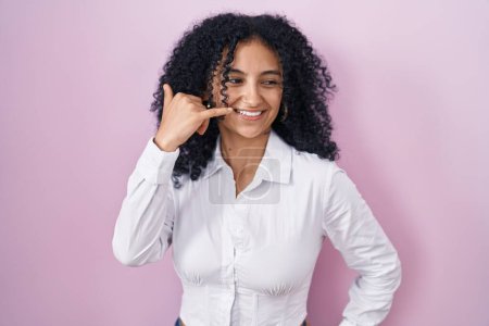 Téléchargez les photos : Femme hispanique avec les cheveux bouclés debout sur fond rose souriant faire geste de téléphone avec la main et les doigts comme parler au téléphone. communiquer les concepts. - en image libre de droit