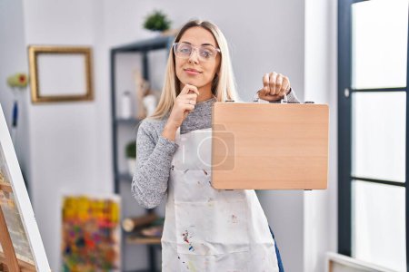 Photo for Young blonde painter woman holding wooden case at art studio serious face thinking about question with hand on chin, thoughtful about confusing idea - Royalty Free Image