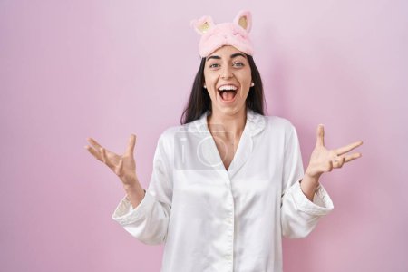 Photo for Young brunette woman wearing sleep mask and pajama celebrating victory with happy smile and winner expression with raised hands - Royalty Free Image