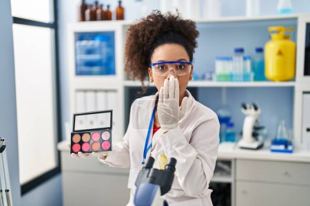 Téléchargez les photos : Young african american woman working at scientist laboratory doing make up covering mouth with hand, shocked and afraid for mistake. surprised expression - en image libre de droit