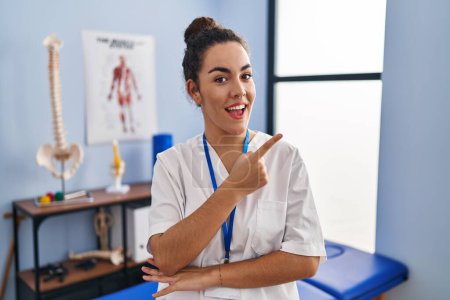 Foto de Young hispanic woman working at rehabilitation clinic with a big smile on face, pointing with hand and finger to the side looking at the camera. - Imagen libre de derechos