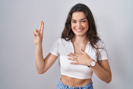 Foto de Young teenager girl standing over white background smiling swearing with hand on chest and fingers up, making a loyalty promise oath - Imagen libre de derechos