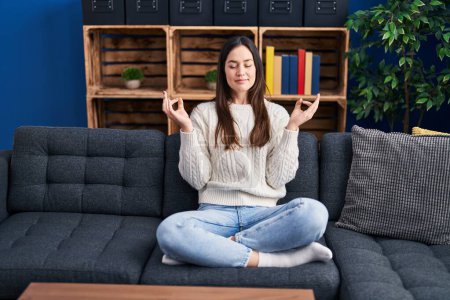 Téléchargez les photos : Jeune femme méditant faire de l'exercice de yoga assis sur le canapé à la maison - en image libre de droit