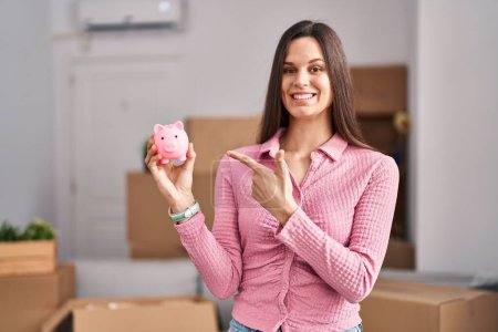 Téléchargez les photos : Young hispanic woman moving to a new home holding piggy bank smiling happy pointing with hand and finger - en image libre de droit