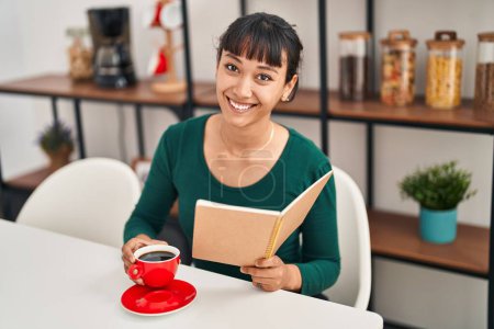 Foto de Young beautiful hispanic woman reading book and drinking coffee sitting on table at home - Imagen libre de derechos