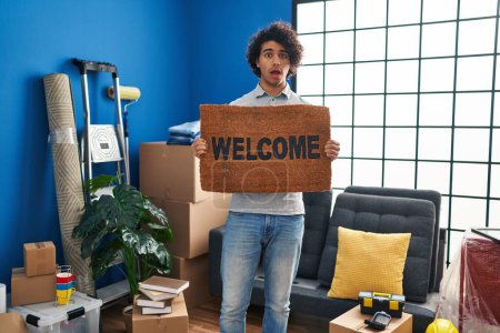Photo for Hispanic man with curly hair holding welcome doormat afraid and shocked with surprise and amazed expression, fear and excited face. - Royalty Free Image