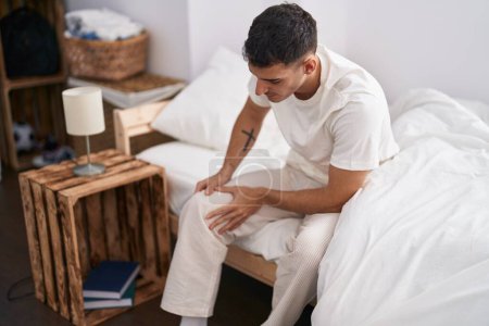 Téléchargez les photos : Young hispanic man suffering for knee injury sitting on bed at bedroom - en image libre de droit