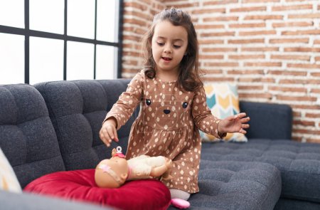 Photo for Adorable hispanic girl playing with baby doll sitting on sofa at home - Royalty Free Image