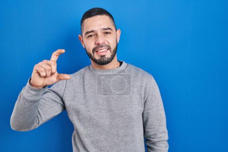 Photo for Hispanic man standing over blue background smiling and confident gesturing with hand doing small size sign with fingers looking and the camera. measure concept. - Royalty Free Image