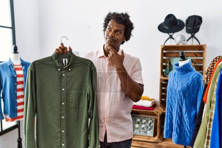Photo for African man with curly hair holding shirt from clothing rack at retail shop serious face thinking about question with hand on chin, thoughtful about confusing idea - Royalty Free Image
