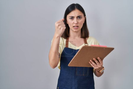 Téléchargez les photos : Hispanic girl wearing apron holding clipboard annoyed and frustrated shouting with anger, yelling crazy with anger and hand raised - en image libre de droit