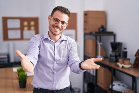 Téléchargez les photos : Jeune homme hispanique au bureau souriant joyeux offrant des mains donnant assistance et acceptation. - en image libre de droit