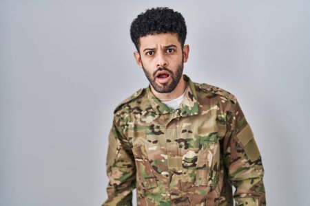 Arab man wearing camouflage army uniform in shock face, looking skeptical and sarcastic, surprised with open mouth 