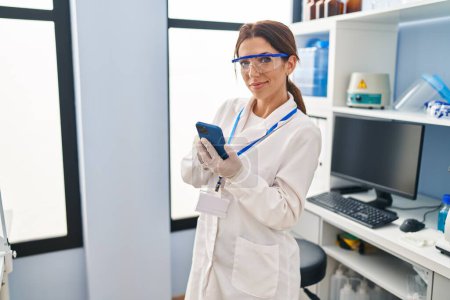 Foto de Mujer hispana joven vistiendo uniforme científico usando smartphone en laboratorio - Imagen libre de derechos