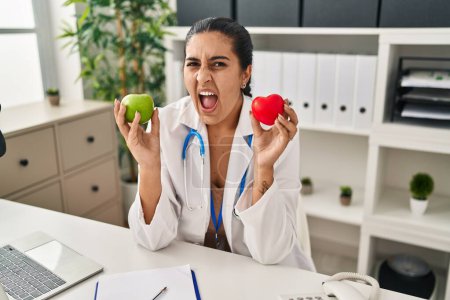 Téléchargez les photos : Young hispanic woman working at dietitian clinic holding green apple angry and mad screaming frustrated and furious, shouting with anger. rage and aggressive concept. - en image libre de droit