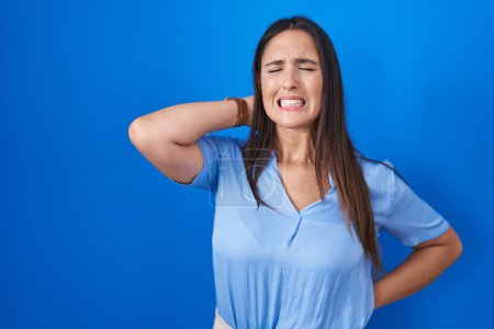 Téléchargez les photos : Jeune femme brune debout sur fond bleu souffrant d'une blessure au cou, toucher le cou avec la main, douleur musculaire - en image libre de droit