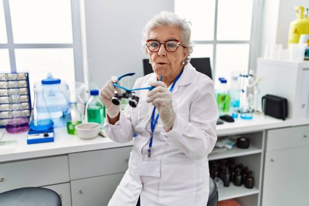 Téléchargez les photos : Femme âgée aux cheveux gris travaillant au laboratoire scientifique à l'aide de loupes faire visage de poisson avec la bouche et les yeux plissés, fou et comique. - en image libre de droit