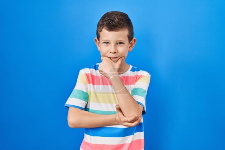 Photo for Young caucasian kid standing over blue background looking confident at the camera smiling with crossed arms and hand raised on chin. thinking positive. - Royalty Free Image