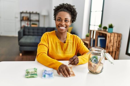Téléchargez les photos : Femme afro-américaine assise sur la table tirant des billets de rand à la maison - en image libre de droit