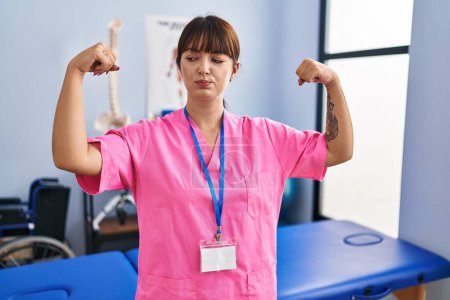 Téléchargez les photos : Young brunette woman working at rehabilitation clinic showing arms muscles smiling proud. fitness concept. - en image libre de droit