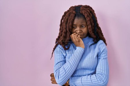 Foto de Mujer africana de pie sobre fondo rosa mirando estresada y nerviosa con las manos en la boca mordiendo las uñas. problema de ansiedad. - Imagen libre de derechos