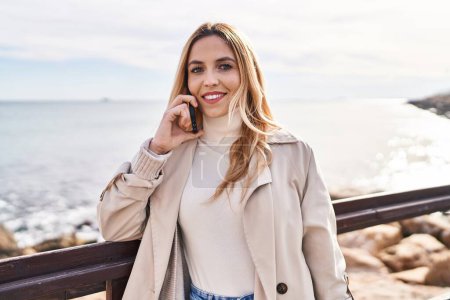 Téléchargez les photos : Jeune femme blonde souriante confiante parlant sur smartphone au bord de la mer - en image libre de droit