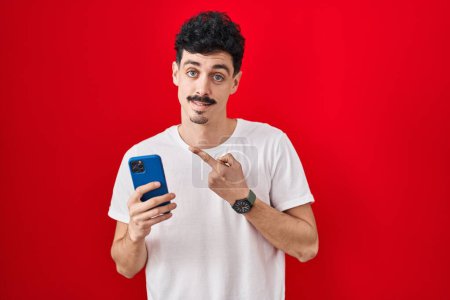 Foto de Hispanic man using smartphone over red background pointing to the back behind with hand and thumbs up, smiling confident - Imagen libre de derechos