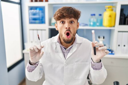 Photo for Arab man with beard working at scientist laboratory holding blood sample afraid and shocked with surprise and amazed expression, fear and excited face. - Royalty Free Image