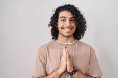 Téléchargez les photos : Hispanic man with curly hair standing over white background praying with hands together asking for forgiveness smiling confident. - en image libre de droit