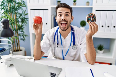 Téléchargez les photos : Young hispanic dietitian man holding doughnut and apple celebrating crazy and amazed for success with open eyes screaming excited. - en image libre de droit