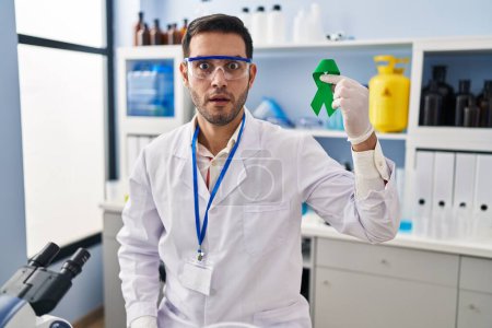 Téléchargez les photos : Jeune homme hispanique avec la barbe travaillant au laboratoire scientifique tenant ruban vert effrayé et étonné avec la bouche ouverte pour surprise, visage incrédule - en image libre de droit