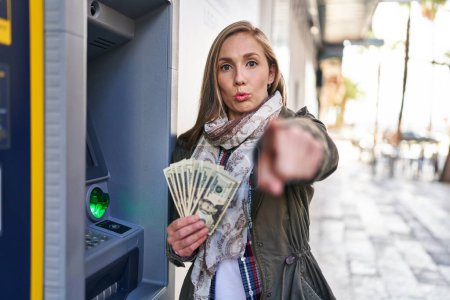 Photo for Young blonde woman holding dollars banknotes from atm machine pointing with finger to the camera and to you, confident gesture looking serious - Royalty Free Image
