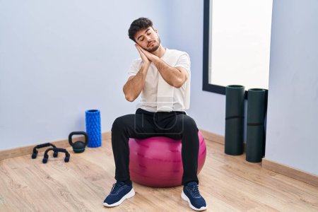 Photo for Hispanic man with beard sitting on pilate balls at yoga room sleeping tired dreaming and posing with hands together while smiling with closed eyes. - Royalty Free Image