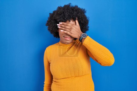 Photo for Black woman with curly hair standing over blue background covering eyes with hand, looking serious and sad. sightless, hiding and rejection concept - Royalty Free Image