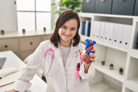 Téléchargez les photos : Femme trisomique portant l'uniforme de médecin tenant le modèle anatomique du coeur à la clinique - en image libre de droit