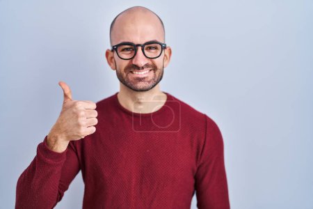 Téléchargez les photos : Jeune homme chauve avec la barbe debout sur fond blanc portant des lunettes faisant des pouces heureux geste avec la main. approuvant l'expression regardant la caméra montrant le succès. - en image libre de droit