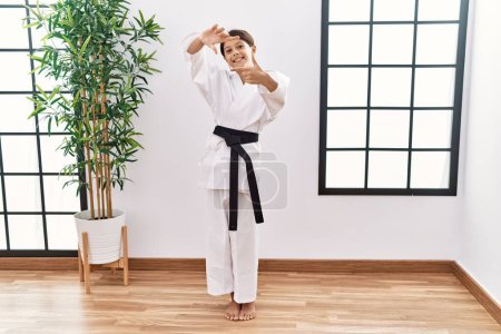 Foto de Joven chica hispana usando kimono de karate y cinturón negro sonriente haciendo marco con manos y dedos con cara feliz. concepto de creatividad y fotografía. - Imagen libre de derechos