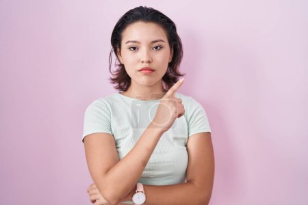Foto de Mujer joven hispana de pie sobre fondo rosa señalando con el dedo de la mano a un lado mostrando publicidad, cara seria y tranquila - Imagen libre de derechos
