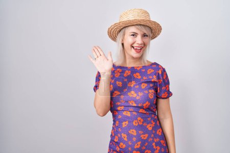 Téléchargez les photos : Jeune femme caucasienne portant des fleurs robe et chapeau d'été renonçant à dire bonjour heureux et souriant, geste de bienvenue amical - en image libre de droit