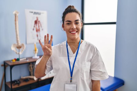 Foto de Young hispanic woman working at rehabilitation clinic showing and pointing up with fingers number four while smiling confident and happy. - Imagen libre de derechos